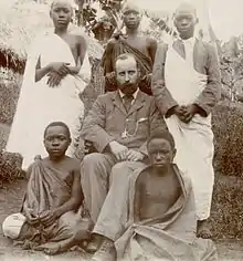 Black and white photo of a seated man wearing a suit suit, surrounded by several young Ugandan men.