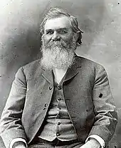 Half-length sitting portrait of man in his fifties with large gray beard and moustache, wearing coat and vest