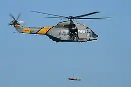 Spanish Air Force Aérospatiale SA330J Puma of 801 Squadron flying in an airshow. It is lifting a stretcher with a hoist. On the side of the helicopter is lettering reading "SAR", in yellow against the military grey color scheme.