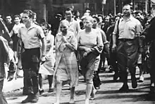 Black-and-white photograph of two women with shaved heads and blank expressions on their face walking down a street in Paris. The women are surrounded by a group of other people, most of whom are smiling.