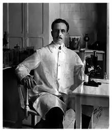 Black and white photo of Charlos Chagas, in his lab coat, sitting next to his microscope and surrounded by flasks and jars