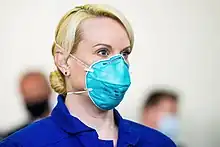 Head-and-shoulders photo of a middle-aged white woman. Her blonde hair is tied in a bun. Her nose and mouth are covered by a teal respirator with head and neck straps. She is wearing royal blue coveralls.