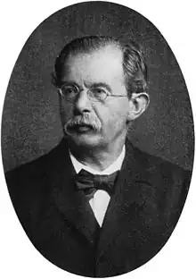 Dignified, ovular, black-and-white, head-and-shoulders portrait of a middle-aged man garbed in a black suit with a bow-tie. The man is bespectacled, with a full mustache and hair which is slicked back, slightly receding in front, and speckled with gray.