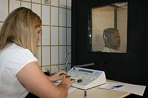 a female medical professional is seated in front of a special sound-proof booth with a glass window, controlling diagnostic test equipment. Inside the booth a middle aged man can be seen wearing headphones and is looking straight ahead of himself, not at the audiologist, and appears to be concentrating on hearing something