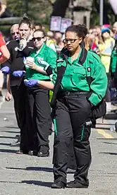 Three St John Ambulance first aiders are standing in unison next to each other. The volunteer on the very left is in a black uniform, the one that was used before 2013, and the other two volunteers on the right are wearing the new uniform.