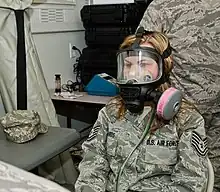 A seated woman wearing a U.S. Air Force combat uniform and a black plastic mask