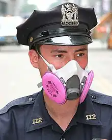 Head-only portrait of a male police officer wearing a navy blue peaked cap emblazoned with the New York City coat of arms and navy uniform shirt with gold collar insignia identifying him as a member of the 112th Precinct. His nose and mouth are covered by a gray rubber respirator with bright pink filters.