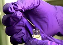 hands wearing purple gloves holding on the right an open vial of smallpox vaccine and on the left a bifurcated needle that has a small droplet dose of the vaccine