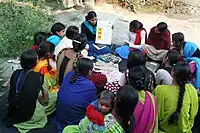  A woman teaches women's literacy class. There is a group of women surrounding a single woman who holds up a board on which there is foreign writing. She is teaching literacy to the group of women.