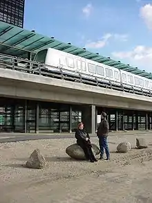 Two people look up at a train stopped at an elevated glass and concrete station