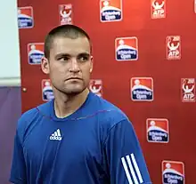 A close to bald man, wearing a blue T-shirts with a white Adidas logo, standing in front of a wall with a dusin St. Petersburg Open logos