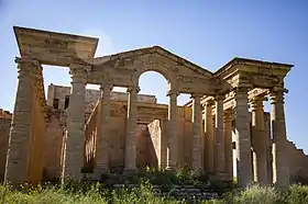 Ruins of a pantheon built with tan stones, with vegetation in the foreground.