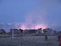 In twilight against snow capped mountains, two firefighters walk through a field flanked by burning rubble of the village