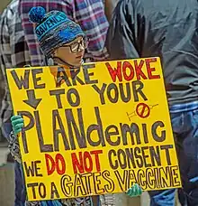 A young adolescent carrying a yellow sign saying, "We are woke to your plandemic. We do not consent to a Gates Vaccine."