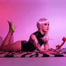 Emily Blue holding red vintage phone, lying down on a black-and-white carpet wearing leather clothing in front of a pink backdrop.