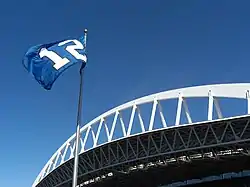 A blue flag with a white number 12 flies against a clear sky. An expansive white roof truss is behind the flagpole.