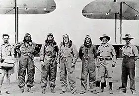 Seven men, four in flying suits, standing in front of the wings of two biplanes