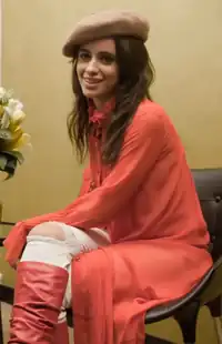 A woman is seen seated on a chair against a beige backdrop as she wears a red outfit and smiles to the camera.