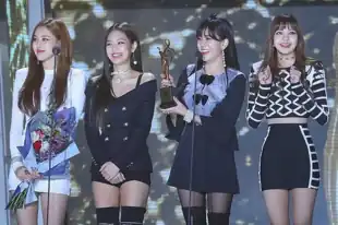 Blackpink standing in a row on stage in front of two microphones. Rose holds a bouquet of flowers while Jisoo holds a trophy.