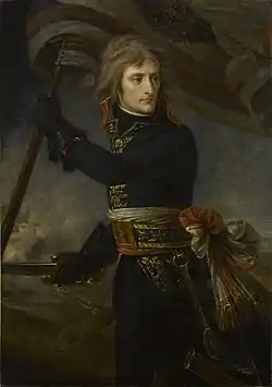 Painting depicts a clean-shaven man with long hair in a dark blue military uniform with a bright sash around his waist. He holds a sword in his right gloved hand and a flag in his left.