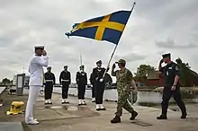 A man in a white naval uniform salutes two men, one in camouflage and one in a blue uniform, who walk towards him while a large blue and yellow Swedish flag is held next to them.
