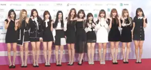 Twelve members of Iz*One pose for a photo in front of a white, pink and purple wall while standing on the red carpet.