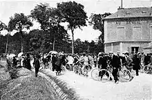 A black-and-white picture of a group of persons with bicycles, standing on a road.