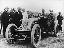 An open-topped sports car, with "3B" written on the front. A driver sits on the right side of the cockpit, and his riding mechanic sits alongside. A group of people crowd around, inspecting the car.