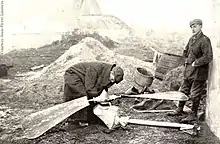 Two men look at a propeller and gear assembly lying on the ground. One is stooping over it, inspecting it closely; the other leans with his back against a whitewashed wall on the right. Buildings are visible in the background.