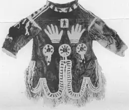 Black and white photograph of a hoodless parka with elaborate designs on the front in contrasting white fur. The designs include a pair of hands and three circles with spokes inside.
