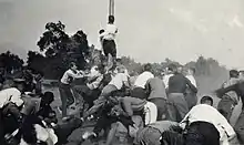 A group of roughly thirty men fight brawling, with some on the ground and one managing to climb a large wooden pole in the center