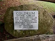 A circular boulder takes up most of the image; behind it can be seen green grass. On the boulder is a light-grey plaque. On this plaque are written the words: "Coldrum Stone Circle has been vested in The National Trust as a Memorial to Benjamin Harrison 1837–1921 of Ightham 10 July 1926". All the letters are in capital letters; those stating "Coldrum Stone Circle" are larger than the others.