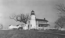 A white lighthouse and attached two-story house on a hill. There is farmland around the house. A large black walnut tree is also pictured behind the station.