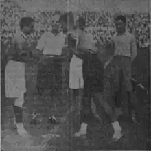 East Bengal captain Paritosh Chakraborty and Mohun Bagan captain Anil Dey shaking hands watched by two men in front of a crowd.