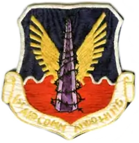 a shield, top half is blue, bottom half red, on it is a grey mololith wrapped in barbed wire with gold eagle wings spread from behind, there is a banner across the bottom with first air commando wing written on it