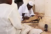 A black man in a cream coloured robe watches a boy in a white robe and white cap write in Arabic on a flat piece of wood. They are both sitting on a tiled floor near a white column.