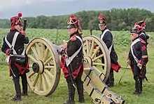 Photo of four reenactors dressed as French artillerymen in dark blue uniforms. They stand near a replica of an early 1800s cannon.