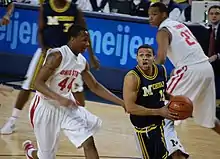 A basketball player in a dark blue uniform drives with the basketball and is defended by a player in a white uniform.