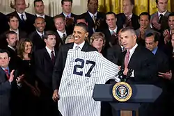 Two men in dark suits stand in front of a crowd in the background and behind a podium in the foreground with the Seal of the President of the United States on it. The two men are holding a white jersey with pinstripes and the number 27 on it.
