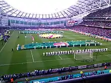 Spectator view of a packed football stadium from behind one of the goals. The opposing stand and part of a lateral stand and the roof structure are seen in the background. A number of people are orderly placed on the pitch, holding banners of several shapes and sizes.