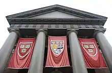 Grand Harvard University building of four pillars in Greek style, with 3 hanging red banners between them. t=The two outer banners show a coat of arms with the word veritas, whilst the central banner has a coat of arms with two roses and an eagle holding a book in addition to the word veritas.