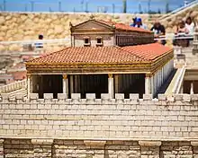 The roof and upper portion of a columned structure rises above a very high stone wall which has wide pilasters decorating the upper part of the wall and a wide square and steps at the foot of the wall.