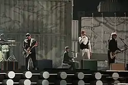 Five men performing on a stage, with four of them playing instruments.