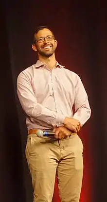 Person with glasses holding a microphone in front of a dark background