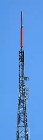 A gray lattice tower, set against a blue sky. A small pink cylindrical antenna and a larger red cylindrical antenna top the structure.
