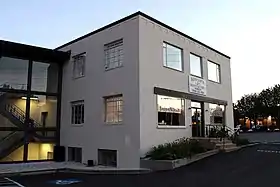 Photograph of a small, rectangular, two story brick building pained white. It is surrounded by an asphalt parking lot. It is annexed to a more modern glass and steel building with an open stairwell showing through the large windows. The sign on the front of the building reads, Kitchen and Bath Factory