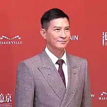 Cheung is facing right and smiling in front of a red background. He is wearing a grey suit.