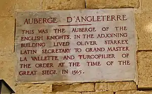 A plaque with the inscription AUBERGE D'ANGLETERRE: THIS WAS THE AUBERGE OF THE ENGLISH KNIGHTS. IN THE ADJOINING BUILDING LIVED OLIVER STARKEY LATIN SECRETARY TO GRAND MASTER VILETTE AND TURCOPILIER OF THE ORDER AT THE TIME OF THE GREAT SIEGE IN 1565
