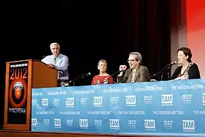 One person standing at a podium and three people sitting at a table with microphones in front of them