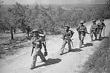 A black and white photograph of men wearing military uniforms marching along a dirt road with a grove of trees in the background